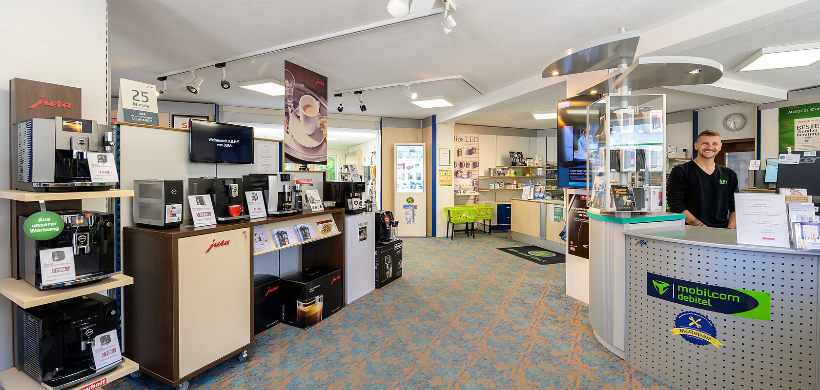Innenansicht eines Geschäfts mit Kaffeemaschinen links und einem Empfangstresen rechts mit einem lächelnden Mitarbeiter.