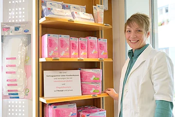 Apothekerin in weißem Kittel steht neben Regal mit pinken Inkontinenzprodukten und Informationsschild in einer Apotheke.