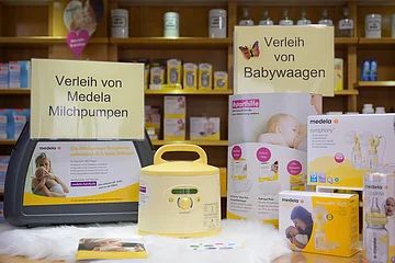 Präsentationstisch in einer Apotheke mit Einer gelben Milchpumpe der Firma "Medela", daneben ein Tragekoffer dazu und ein Schild mit dem Schriftzug "Verleih von Medela Milchpumpen". Ein weiteres Schild mit dem Schriftzug "Verleih von Babywaagen" rechts daneben, mit Produkten und Informationsmaterial auf einem Tisch.