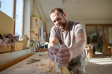 Mann hobelt Holz auf Werkbank in heller Werkstatt mit Werkzeug vor einem Fenster.