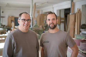 Zwei Männer in grauen Oberteilen stehen in einer Werkstatt mit Holzbearbeitungsmaschinen.