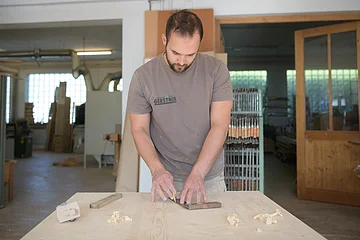 Mann arbeitet mit Holz und Lineal an einem großen Holztisch in einer Werkstatt.