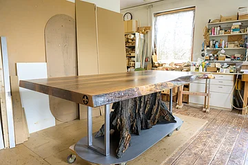 Dunkler Holztisch mit Baumstamm als Tischbein in Werkstatt mit Werkzeugen und Holzplatten im Hintergrund.