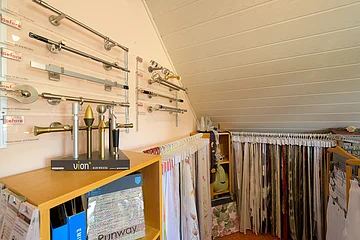 Raum mit Stoffmustern an Kleiderbügeln und verschiedenen Gardinenstangen an der Wand unter einer schrägen Decke.
