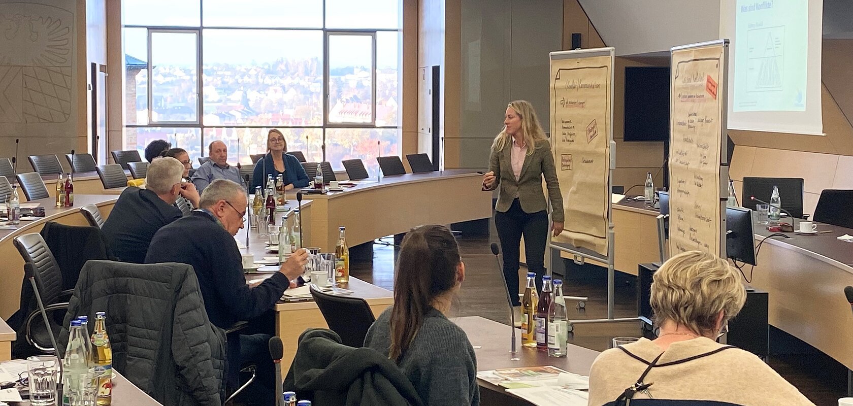 Seminarraum mit hellen Tischen und schwarzen Stühlen, mehreren Personen sitzen an den Tischen, eine Frau präsentiert vor Flipcharts und einem Projektorbild.