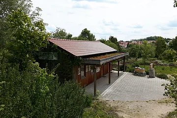 Holzgebäude mit rotem Dach und begrüntem Vordach, umgeben von Bäumen und einem gepflasterten Vorplatz, bei bewölktem Himmel.