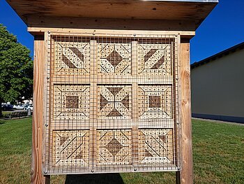 Insektenhotel aus Holz mit neun quadratischen Fächern, jedes mit verschiedenen Mustern und Löchern, im Freien auf einer grünen Rasenfläche mit blauem Himmel im Hintergrund.