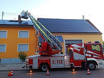 Feuerwehrfahrzeug das auf der Straße steht mit ausgefahrener Drehleiter und Personen oben auf der Drehleiter, vor einem orange gestrichenem Haus mit Solarpanelen auf dem Dach.