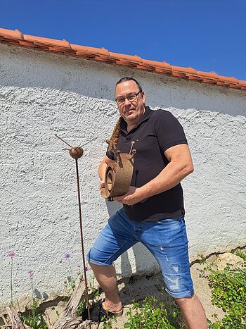 Mann mit Brille und Zopf hält eine große Schneckenfigur aus rostigem Metall vor einer weißen Wand mit rotem Dach.