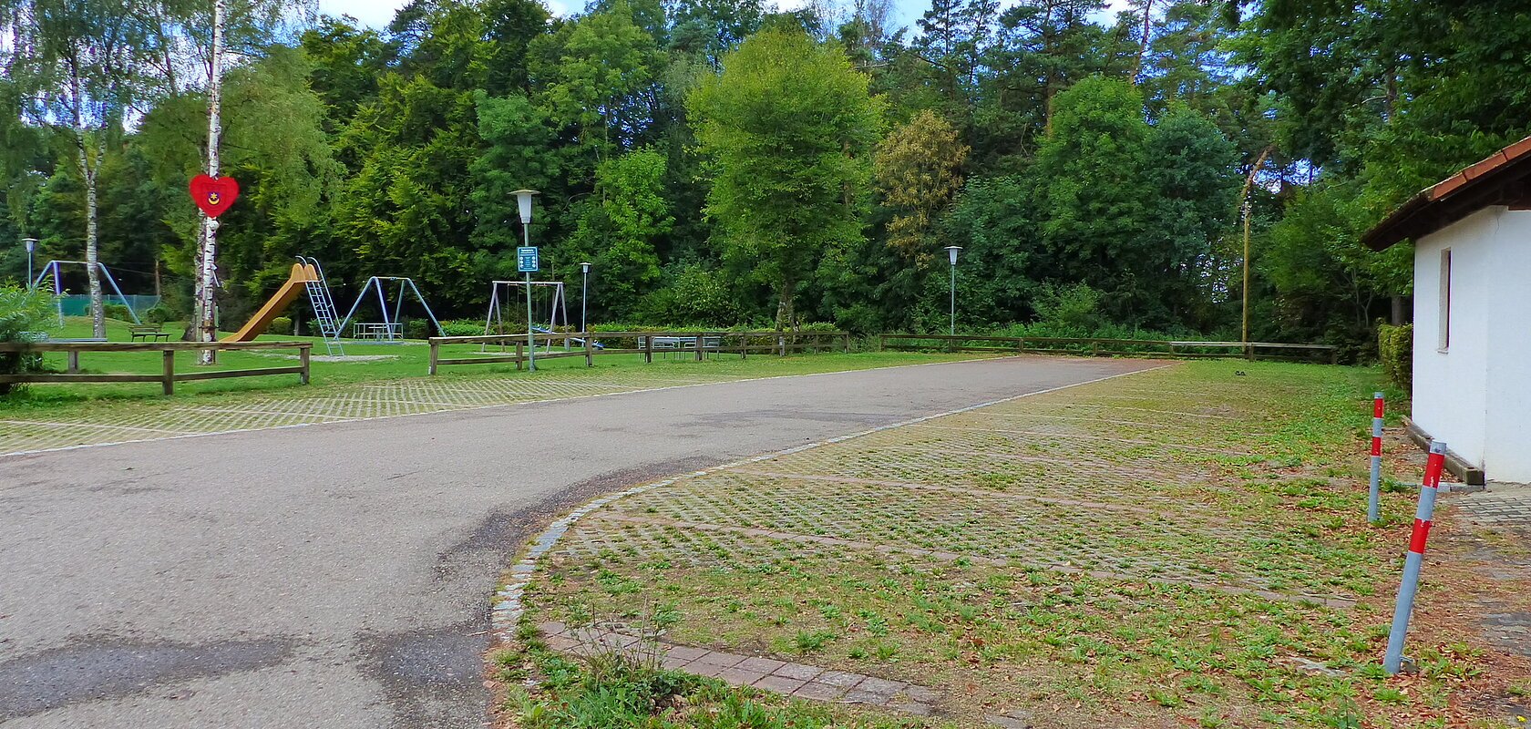 Parkplatz mit Grünfläche rechts im Bild und einem Spielplatz mit Rutsche und Schaukeln vor Wald im Hintergrund. Ein Maibaum mit Herz steht auf der Grünfläche links.