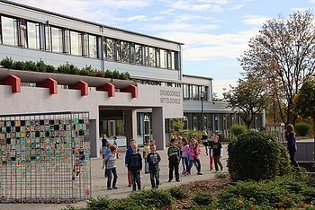 Graues Schulgebäude mit flachem Dach, mit Kindern auf dem Schulhof und einem Kunstwerk aus bemalten Kacheln an einem Gitter links im Bild. Rechts im Bild bepflanzte Fläche mit Büschen und Pflanzen, sowie Bäumen im Hintergrund bei bewölktem Himmel.