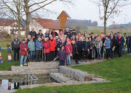 Große Gruppe älterer Menschen steht im Park vor einem kleinen Wasserbecken mit Treppe und Geländer. Monatliche Wanderung für Senioren vom Verein Soziales Miteinander