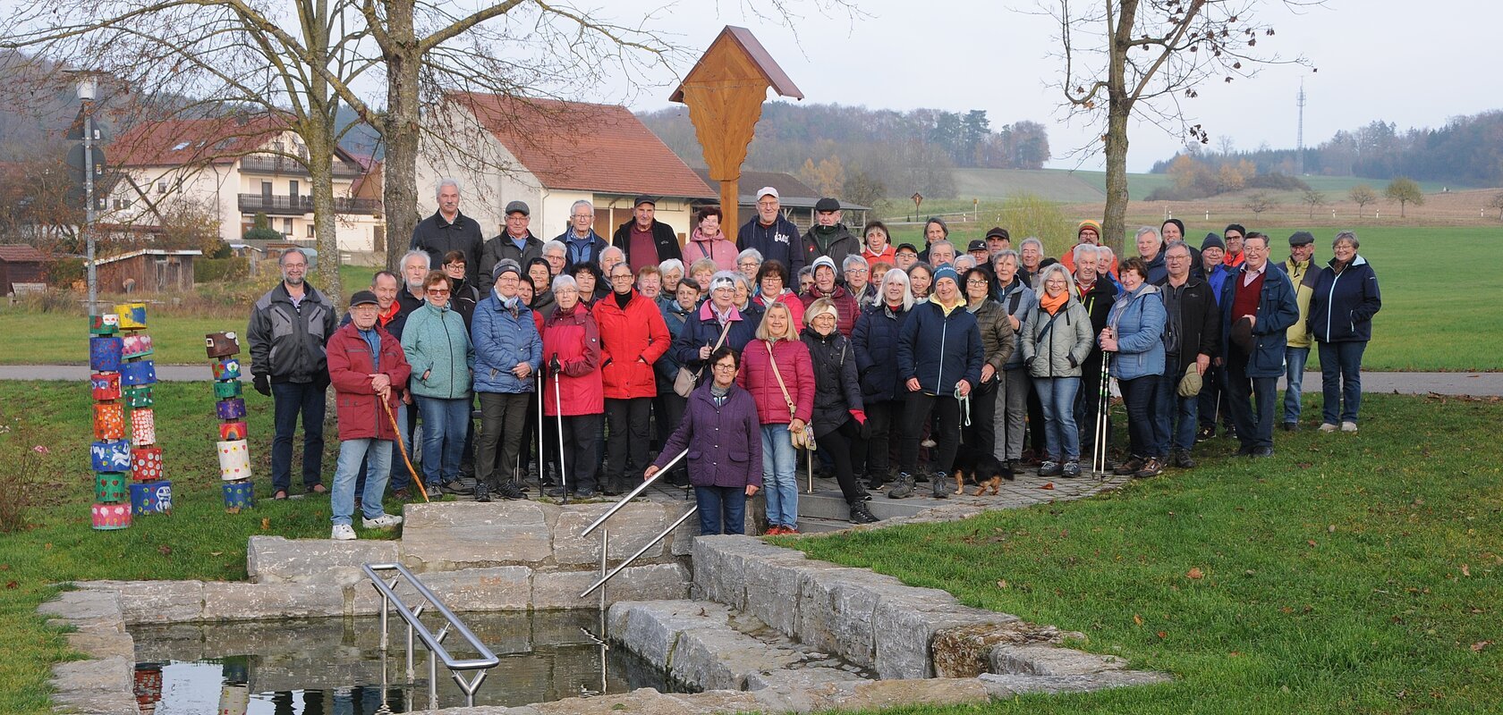 Große Gruppe älterer Menschen steht im Park vor einem kleinen Wasserbecken mit Treppe und Geländer. Monatliche Wanderung für Senioren vom Verein Soziales Miteinander