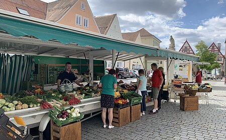Marktstand des Wochenmarkts in Monheim mit Obst und Gemüse in der Auslage auf dem gepflastertem Marktplatz. Mehrere Personen kaufen dort ein. Fachwerkhäuser im Hintergrund und bewölkter Himmel.