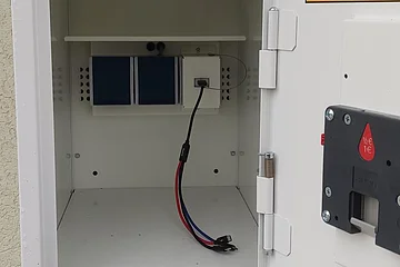 Offener weißer Metallkasten, eine E-Bike Ladestation zeigt die Innenausstattung mit Kabeln und Steckdosen an einer Außenwand stehend, oberste Tür ist geöffnet.