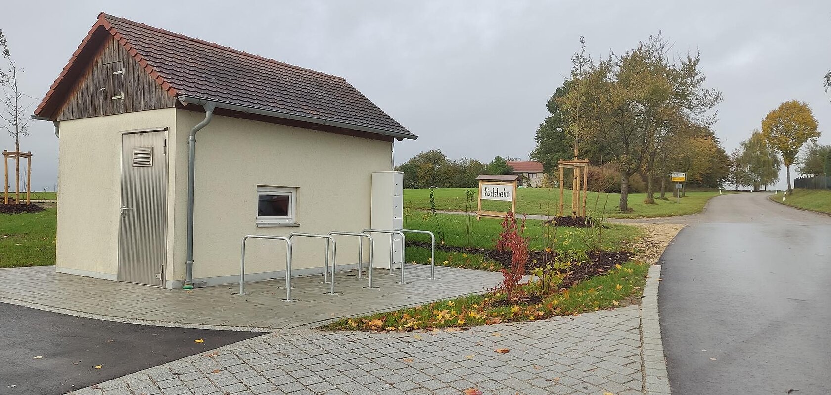Kleines Gebäude mit Metalltür und kleinem Fenster, sowie fünf Fahrradständern auf gepflastertem Platz neben einer Straße und einem Schild mit Ortsnamen Flotzheim, unter bewölktem Himmel.