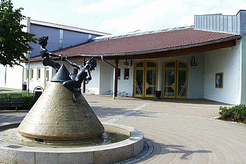 Kegelförmiger Steinbrunnen mit Wasser gefüllt, zwei spielende Kinderfiguren aus Metall oben drauf vor einem Gebäude mit gelben Türen und rotem Dach, auf einem gepflastertem Gebäude. Links im Bild ein weiteres angrenzendes hohes Gebäude bei blauem Himmel.