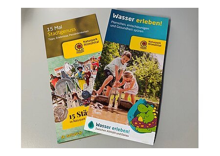 Zwei Broschüren des Naturparks Altmühltal zu Stadtgenuss und Wassererlebnissen mit Bildern und Texten liegen übereinander auf einem weißen Tisch.