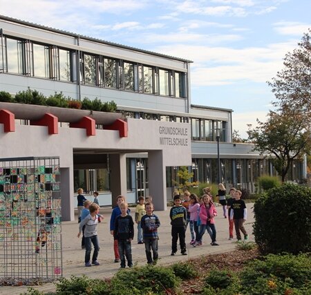 Graues Schulgebäude mit flachem Dach, mit Kindern auf dem Schulhof und einem Kunstwerk aus bemalten Kacheln an einem Gitter links im Bild. Rechts im Bild bepflanzte Fläche mit Büschen und Pflanzen, sowie Bäumen im Hintergrund bei bewölktem Himmel.
