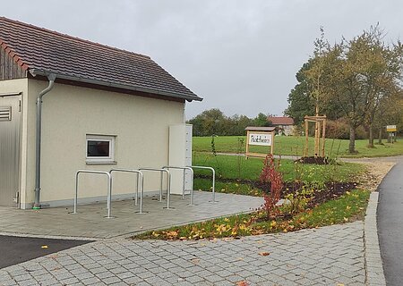Kleines Gebäude mit Metalltür und kleinem Fenster, sowie fünf Fahrradständern auf gepflastertem Platz neben einer Straße und einem Schild mit Ortsnamen Flotzheim, unter bewölktem Himmel.