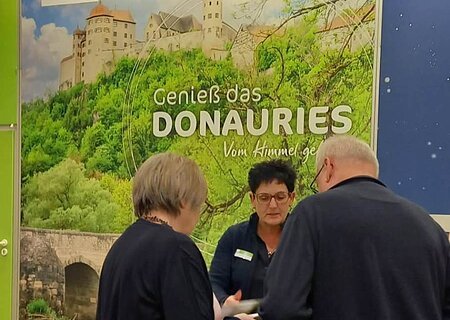 Drei Personen an einem Messestand mit Werbeplakat für Donauries Ferienland im Hintergrund und Losbox auf dem Boden.