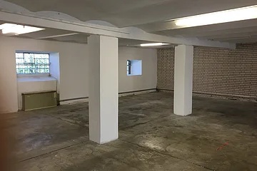 Leerer Raum mit zwei weißen Stützsäulen, Betonboden und kleinen Fenstern mit Gitter.