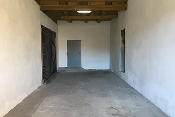 Leerer Raum mit Betonboden, weißen Wänden, Holzbalkendecke, grauer Tür am Ende und einem Durchgang rechts.