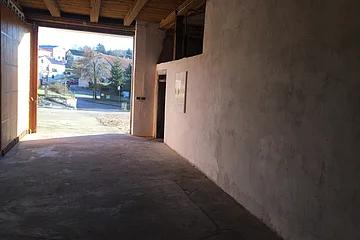 Blick aus einem dunklen Raum nach draußen auf eine helle Straße mit Häusern und Bäumen im Hintergrund. Oben eine Holzdecke mit Holzbalken.