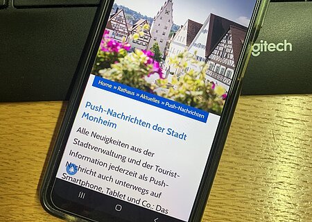 Schwarzes Smartphone zeigt Webseite der Stadt Monheim mit aufgerufener Seite zu Push-Nachrichten. Das Smartphone liegt auf der Tastatur und Holzoberfläche.