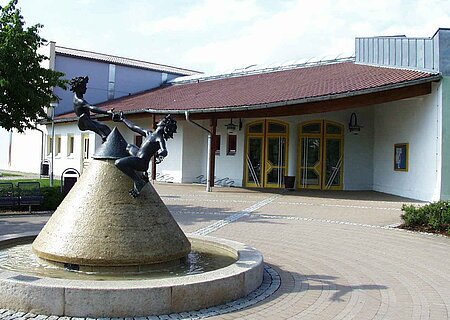 Kegelförmiger Steinbrunnen mit Wasser gefüllt, zwei spielende Kinderfiguren aus Metall oben drauf vor einem Gebäude mit gelben Türen und rotem Dach, auf einem gepflastertem Gebäude. Links im Bild ein weiteres angrenzendes hohes Gebäude bei blauem Himmel.