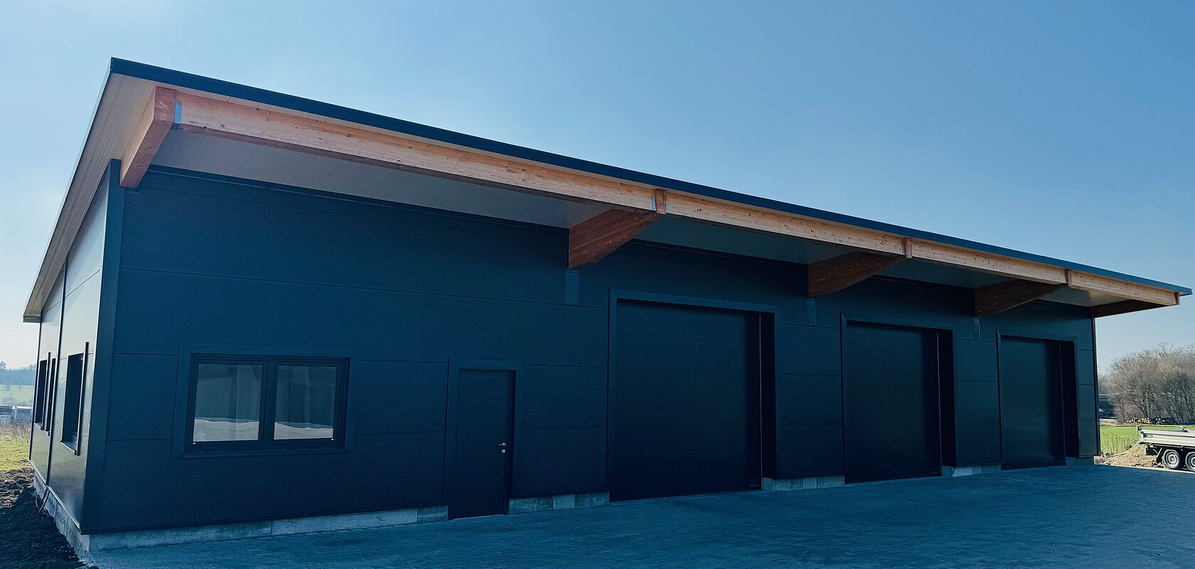 Schwarzes Industriegebäude mit drei Toren, einer Tür und einem Fenster an der Front, unter blauem Himmel an gepflastertem Boden.
