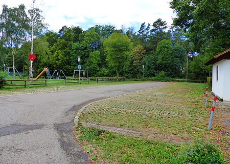 Parkplatz mit Grünfläche rechts im Bild und einem Spielplatz mit Rutsche und Schaukeln vor Wald im Hintergrund. Ein Maibaum mit Herz steht auf der Grünfläche links.