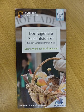 Auf einem Holztisch die Broschüre des regionalen Einkaufsführers aus dem Landkreis Donau-Ries vom Herausgeber Ferienland Donau-Ries und deren zwei Logos auf dem Cover. Auf dem Foto ein Oberarm einer Person, die ein Flechtkorb voller Lebensmittel trägt mit unscharfen Hintergrund. Darüber mittig der Titel "Der regionale Einkaufsführer".
