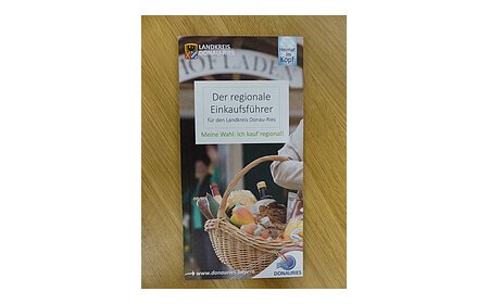Auf einem Holztisch die Broschüre des regionalen Einkaufsführers aus dem Landkreis Donau-Ries vom Herausgeber Ferienland Donau-Ries und deren zwei Logos auf dem Cover. Auf dem Foto ein Oberarm einer Person, die ein Flechtkorb voller Lebensmittel trägt mit unscharfen Hintergrund. Darüber mittig der Titel "Der regionale Einkaufsführer".