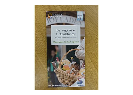 Auf einem Holztisch die Broschüre des regionalen Einkaufsführers aus dem Landkreis Donau-Ries vom Herausgeber Ferienland Donau-Ries und deren zwei Logos auf dem Cover. Auf dem Foto ein Oberarm einer Person, die ein Flechtkorb voller Lebensmittel trägt mit unscharfen Hintergrund. Darüber mittig der Titel "Der regionale Einkaufsführer".