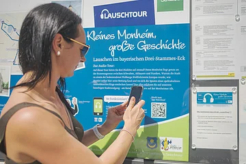Frau mit Smartphone steht vor Infotafel zur Lauschtour in Monheim, Bayern, mit dem Titel "Kleines Monheim, große Geschichte" bei Sonnenschein. Frau hält ihr Smartphone an einen QR-Code.