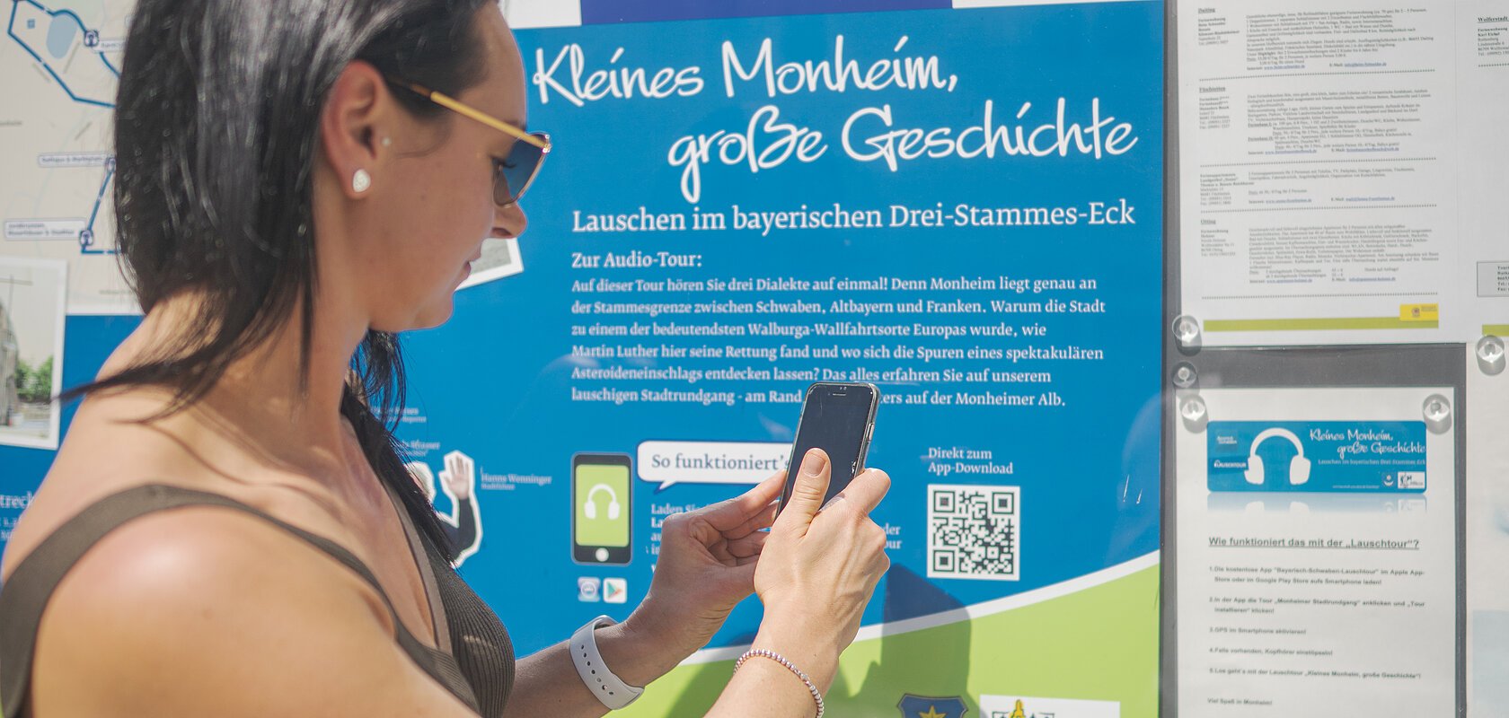 Frau mit Smartphone steht vor Infotafel zur Lauschtour in Monheim, Bayern, mit dem Titel "Kleines Monheim, große Geschichte" bei Sonnenschein. Frau hält ihr Smartphone an einen QR-Code.