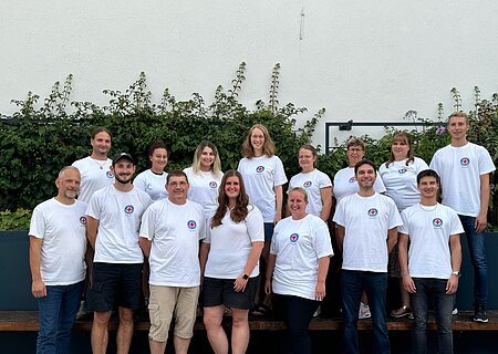 Gruppenfoto von 15 Personen in weißen T-Shirts mit dem Logo der Wasserwacht drauf, stehen blickend in die Kamera vor einer weißen Wand mit Pflanzen im Hintergrund.