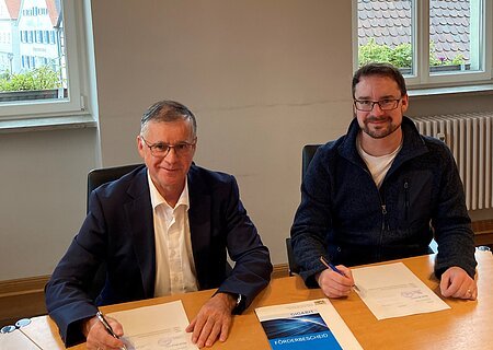 Zwei Männer blickend in die Kamera sitzen an einem Holztisch und unterschreiben Dokumente. Auf dem Tisch liegt eine Mappe mit der Aufschrift „GIGABIT FÖRDERBESCHEID“.