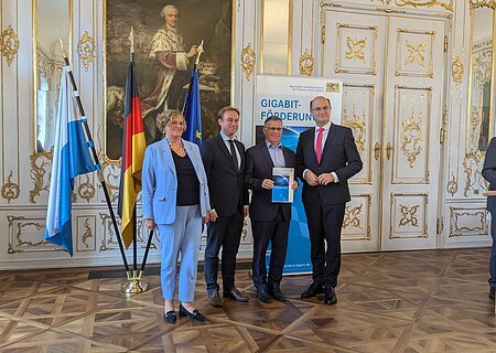 Fünf Personen in Anzügen stehen vor drei Fahnen und einem Banner mit der Aufschrift „Gigabit-Förderung“ in einem prunkvollen Raum. Rechts im Bild steht ein weiterer Mann im Anzug am Rednerpult.