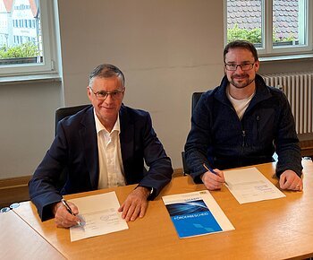 Zwei Männer blickend in die Kamera sitzen an einem Holztisch und unterschreiben Dokumente. Auf dem Tisch liegt eine Mappe mit der Aufschrift „GIGABIT FÖRDERBESCHEID“.