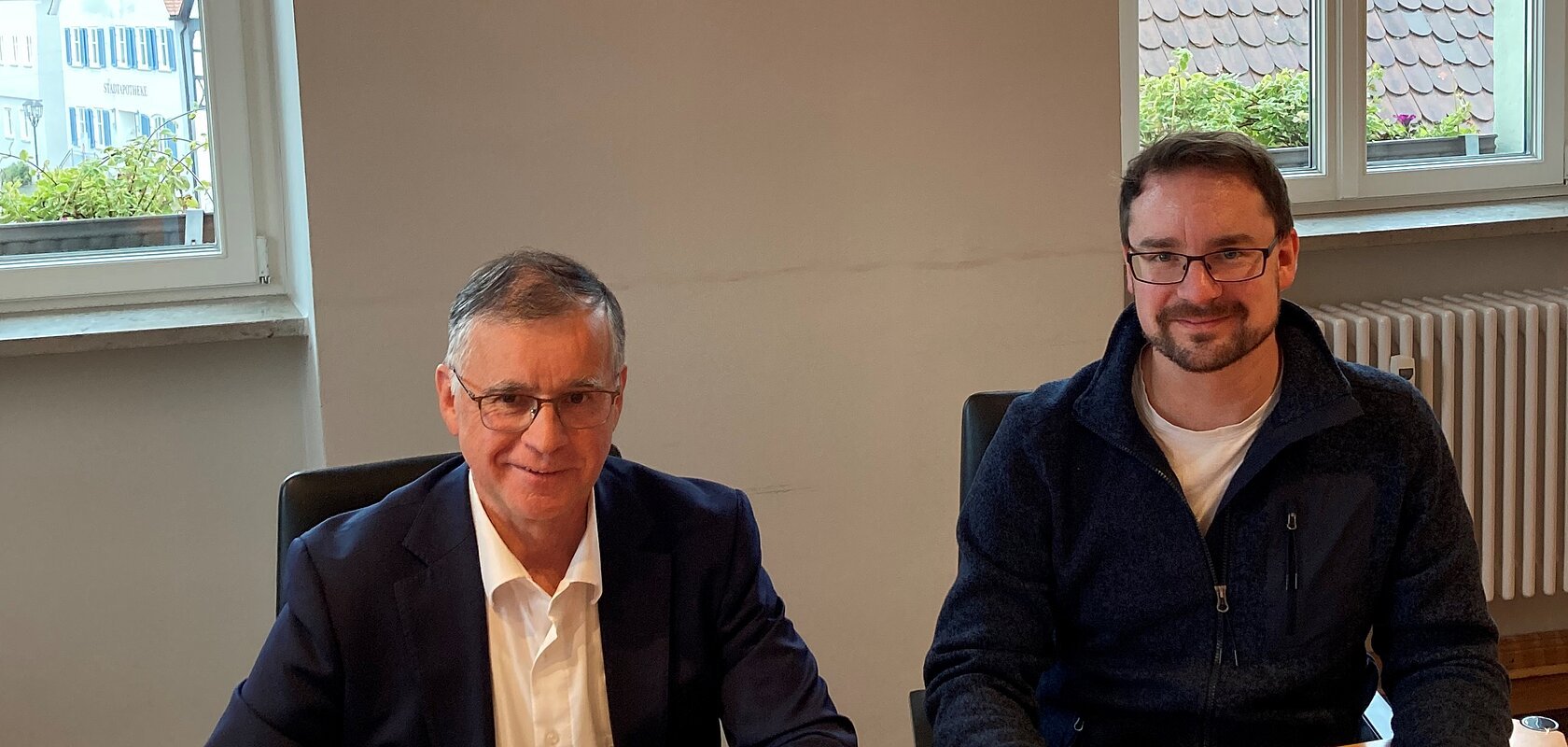 Zwei Männer blickend in die Kamera sitzen an einem Holztisch und unterschreiben Dokumente. Auf dem Tisch liegt eine Mappe mit der Aufschrift „GIGABIT FÖRDERBESCHEID“.