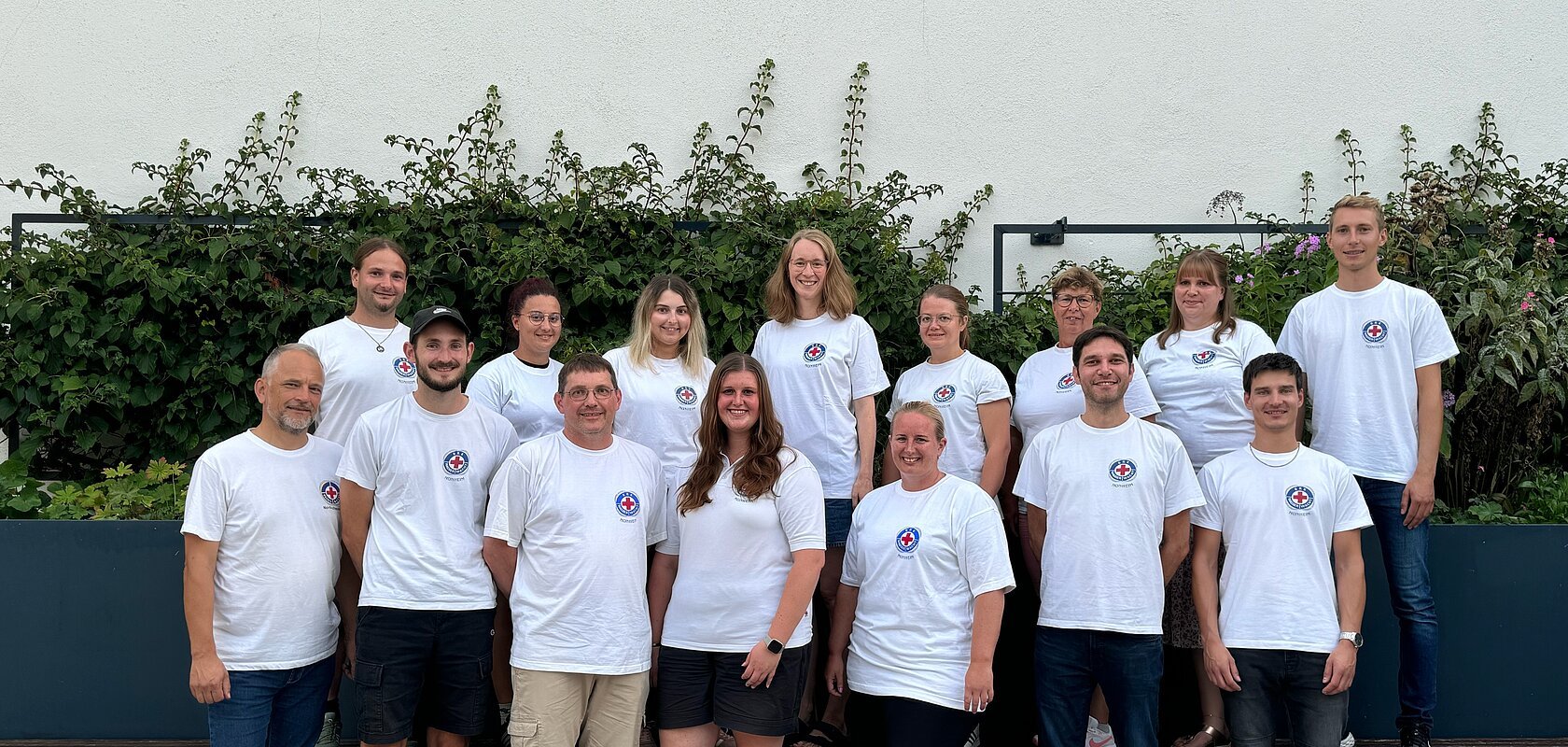Gruppenfoto von 15 Personen in weißen T-Shirts mit dem Logo der Wasserwacht drauf, stehen blickend in die Kamera vor einer weißen Wand mit Pflanzen im Hintergrund.