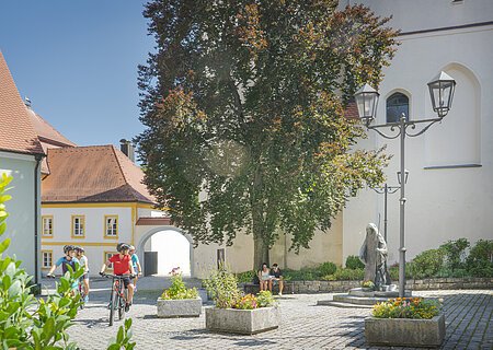 Innenhof mit Radfahrern, Baum, Sitzbank, Statue und historischen Gebäuden bei Sonnenschein