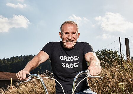 Mann mit schwarzem T-Shirt mit der weißen Aufschrift "DREGG-SAGG" und Jeans sitzt lachend auf orangefarbenem Fahrrad auf Feldweg bei Sonnenschein