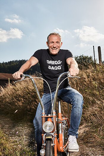 Mann mit schwarzem T-Shirt mit der weißen Aufschrift "DREGG-SAGG" und Jeans sitzt lachend auf orangefarbenem Fahrrad auf Feldweg bei Sonnenschein