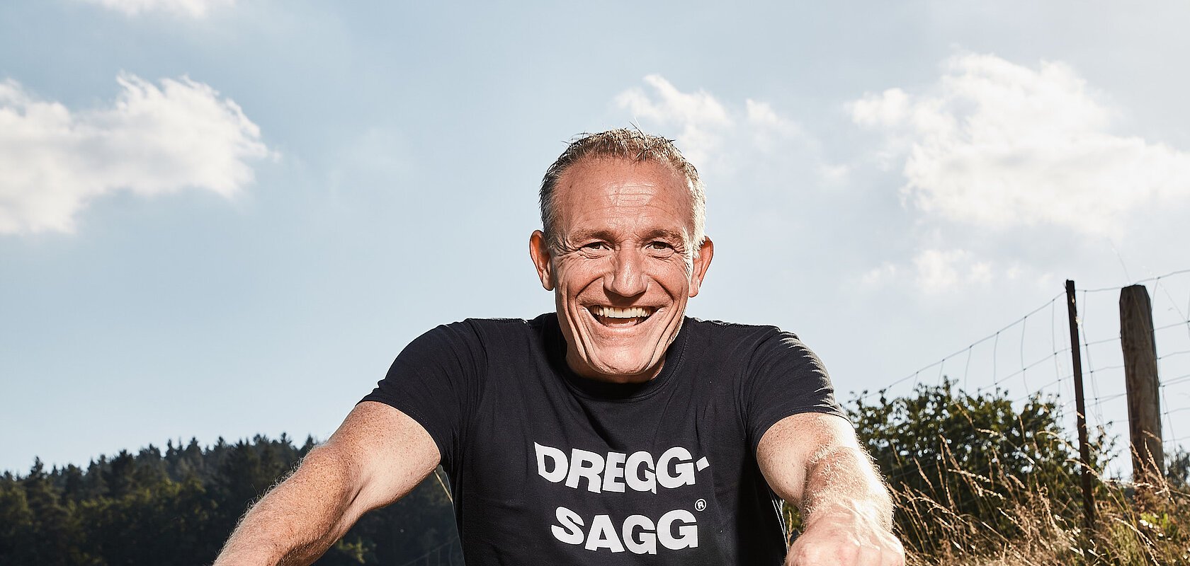 Mann mit schwarzem T-Shirt mit der weißen Aufschrift "DREGG-SAGG" und Jeans sitzt lachend auf orangefarbenem Fahrrad auf Feldweg bei Sonnenschein