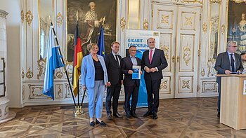 Fünf Personen in Anzügen stehen vor drei Fahnen und einem Banner mit der Aufschrift „Gigabit-Förderung“ in einem prunkvollen Raum. Rechts im Bild steht ein weiterer Mann im Anzug am Rednerpult.