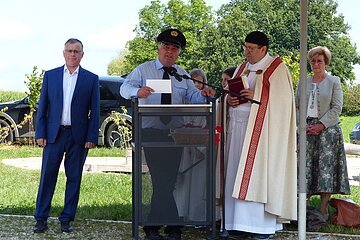 Im Vordergrund drei Personen bei einer Veranstaltung im Freien vor einer Wiese. Darunter ein Mann in Uniform sprechend am Rednerpult und ein Geistlicher mit Buch rechts daneben, links vom Rednerpult steht ein Mann in blauem Anzug. Im Hintergrund weitere Personen, sowie Bäume und ein dunkles geparktes Auto zu sehen.