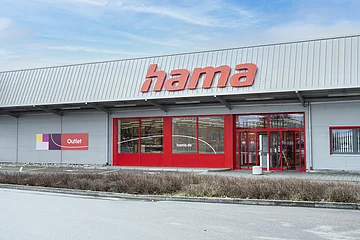 Graue Fassade eines Outlet-Geschäfts mit rotem Schriftzug "Hama", roten Eingangstüren und roten Fensterumrandungen, bei bewölktem Himmel. Vor dem Geschäft eine Hecke entlang einer Straße.